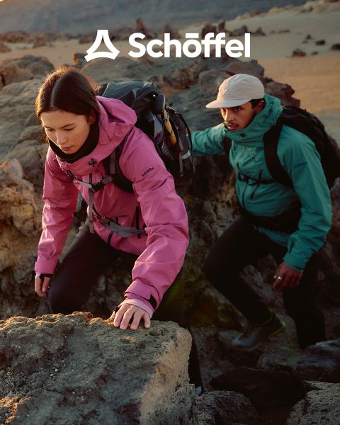 Due persone con abbigliamento da outdoor camminano su una collina erbosa con montagne e nuvole sullo sfondo. Sopra di loro c'è il logo Schöffel, sotto una linea ondulata verde decorativa.