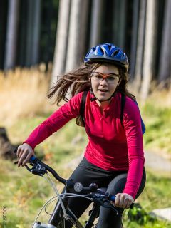 Una donna che indossa una camicia rossa a maniche lunghe, un casco e degli occhiali sta andando in bicicletta in una zona erbosa e boschiva in una giornata di sole.