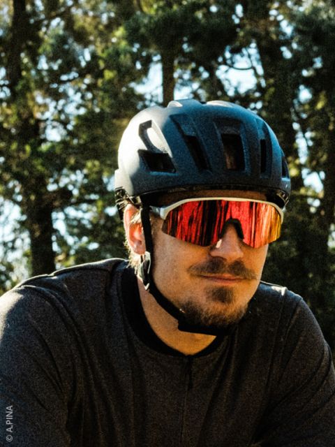 Un uomo con un casco da bici nero, occhiali da sole rossi riflettenti e una camicia scura è in piedi all'aperto con degli alberi sullo sfondo.