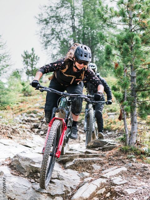 Due ciclisti con casco stanno percorrendo un sentiero roccioso e boscoso con le loro mountain bike, concentrati e destreggiandosi tra alberi e terreni accidentati.