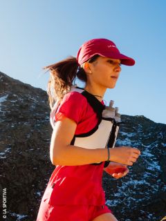 Una donna in abbigliamento da corsa rosso fa jogging con un gilet da idratazione sotto un cielo blu su un percorso di montagna.