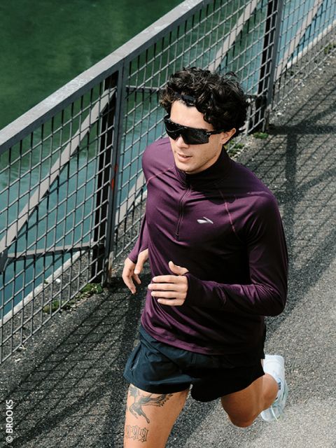 Una persona che indossa occhiali da sole e una maglia a maniche lunghe viola cammina all'aperto accanto a una recinzione metallica su un sentiero lastricato.