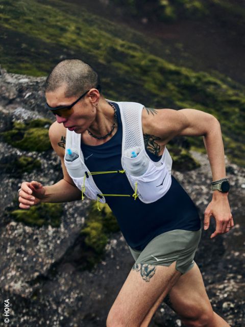 Una persona con tatuaggi cammina su un sentiero roccioso e muschioso in salita, indossa occhiali da sole, un gilet da idratazione e uno smartwatch.