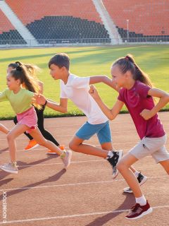 Quattro bambini corrono su una pista in uno stadio, sorridono e si godono la corsa in una giornata di sole.
