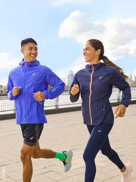 Due persone in abbigliamento sportivo fanno jogging all'aperto su un sentiero lastricato lungo un fiume e si sorridono, mentre sullo sfondo si vedono lo skyline della città e il cielo azzurro.