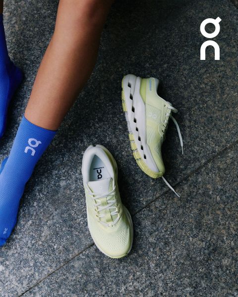 Una persona con calzini azzurri accanto a un paio di scarpe da corsa ON giallo chiaro su un pavimento di pietra grigia, con il logo ON nell'angolo in alto a destra.