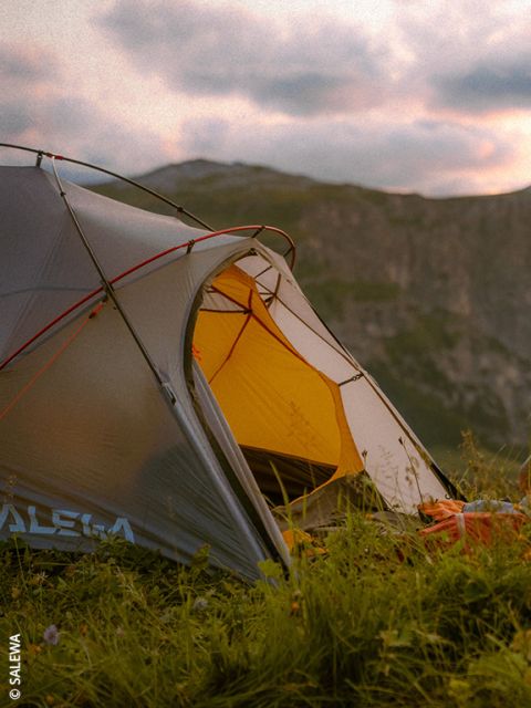 Una tenda grigia e gialla montata in un prato al tramonto con le montagne sullo sfondo.