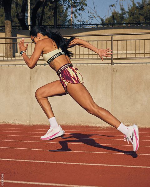 Una donna in abbigliamento sportivo scatta su una pista rossa all'aperto, le braccia che pompano e il corpo che si piega in avanti a metà passo.