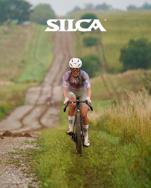 Ciclista che pedala in salita su una strada sterrata attraverso campi erbosi. Indossa un casco e abbigliamento da ciclismo, con il logo "SILCA" nella parte superiore dell'immagine.