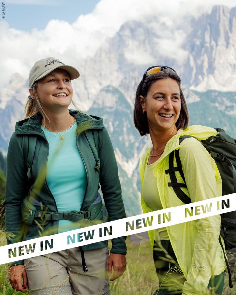 Due donne con attrezzatura da trekking sorridono mentre sono in piedi di fronte a una catena montuosa, con il testo "NEW IN" nella parte inferiore dell'immagine.