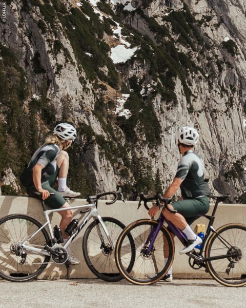 Due ciclisti in abbigliamento coordinato si fermano a una sbarra stradale di montagna, con scogliere rocciose e chiazze di neve sullo sfondo.