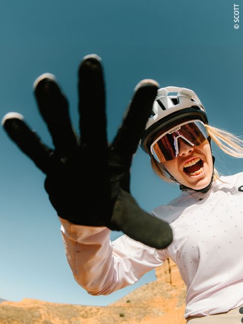 Un ciclista con casco e occhiali da sole sorride energicamente e allunga una mano guantata verso la fotocamera davanti a un cielo azzurro limpido.