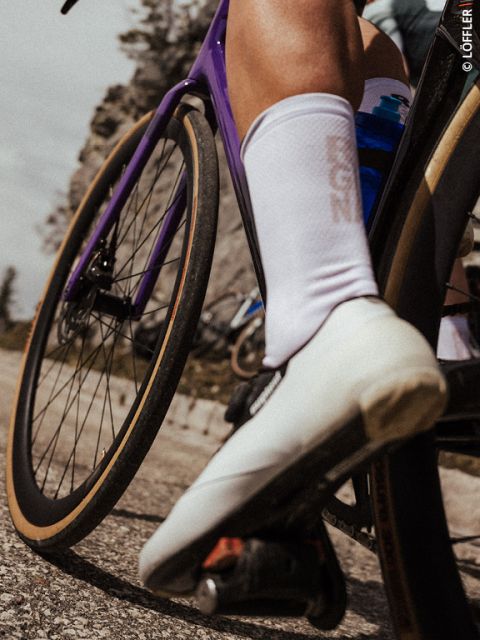 Primo piano della gamba e della scarpa bianca di un ciclista su una bicicletta viola, con una strada sfocata e un paesaggio roccioso sullo sfondo.