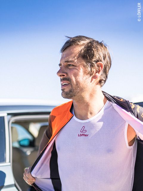 Un uomo all'aperto, che indossa una maglia a rete bianca e una giacca arancione, è in piedi accanto a un'auto sotto un cielo azzurro limpido.