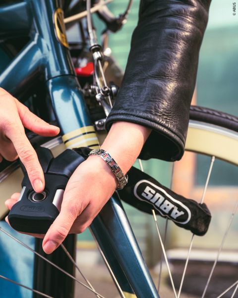 Una persona con una giacca di pelle chiude una bicicletta blu con un lucchetto per bici ABUS, concentrandosi sulla protezione del telaio e della ruota.