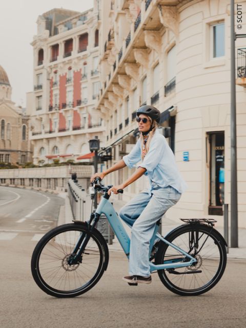 Una donna con casco e occhiali da sole guida una bicicletta blu lungo una strada fiancheggiata da edifici eleganti.