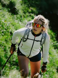 Una donna cammina in salita su un sentiero verde, indossa occhiali da sole, una giacca bianca, uno zaino e usa bastoncini da trekking.