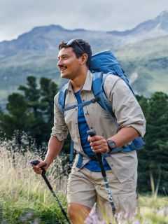 Un uomo sorridente con i capelli lunghi e la barba, che indossa una camicia blu e uno zaino, è in piedi all'aperto di fronte a uno sfondo naturale sfocato.