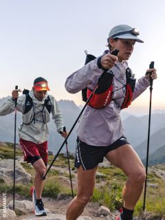 Due escursionisti con lampade frontali e bastoncini da trekking scalano un sentiero di montagna roccioso all'alba, in abbigliamento sportivo e con zaini, con le montagne sullo sfondo.