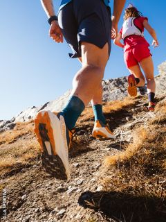 Due trail runner, visti da dietro da una angolazione bassa, corrono in salita su un sentiero roccioso sotto un cielo azzurro limpido.