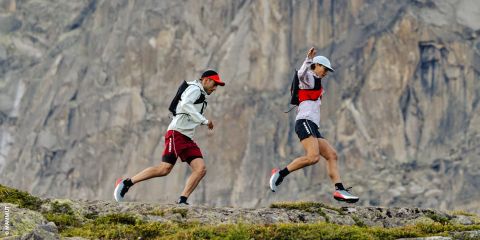 Due persone che fanno trail running su terreno roccioso con una grande parete rocciosa sullo sfondo, entrambe con zaini, cappelli e abbigliamento sportivo.