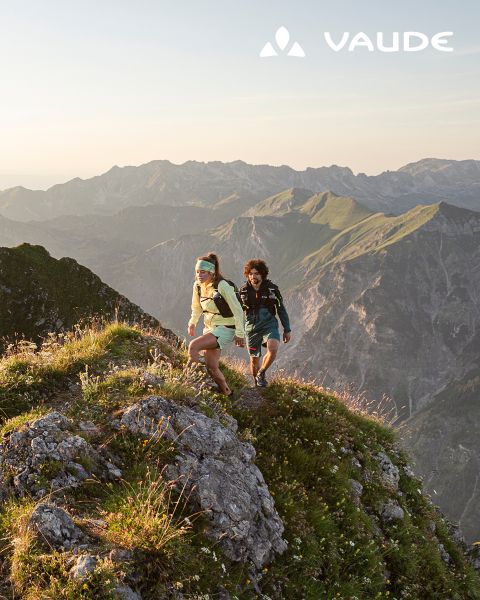 Due escursionisti con zaini in spalla si arrampicano su una cresta erbosa all'alba, con montagne scoscese sullo sfondo. Il logo Vaude appare nell'angolo.