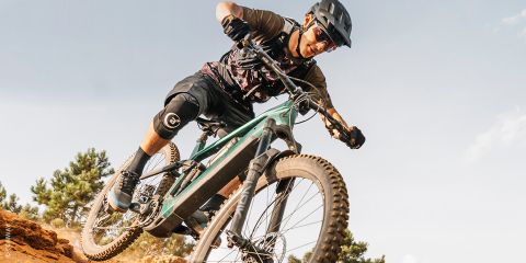 Un ciclista con equipaggiamento protettivo percorre un sentiero sterrato con la sua mountain bike in una giornata di sole, inclinandosi bruscamente in una curva.