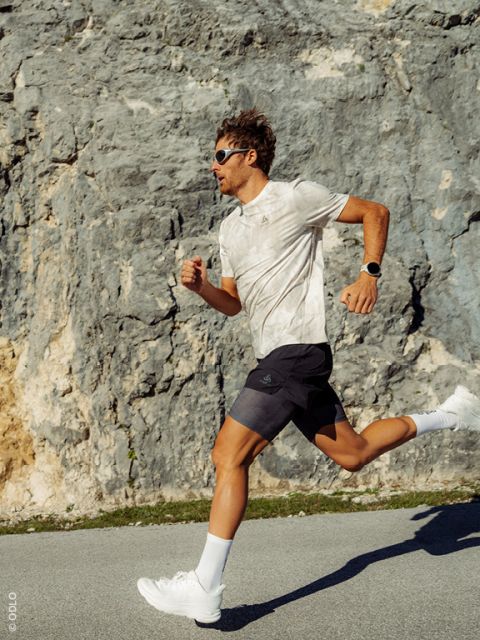 Un uomo con occhiali da sole e abbigliamento sportivo cammina lungo una strada che costeggia una parete rocciosa, sotto la luce intensa del sole.