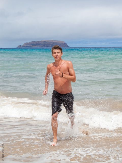 Ein hemdsärmeliger Mann mit Tattoos und schwarzen Badeshorts steht bei Sonnenuntergang im flachen Meerwasser und schaut in die Kamera. Hinter ihm rollen die Wellen heran.