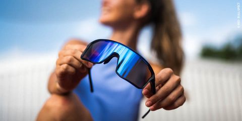 Una donna con un top blu tiene in mano occhiali da sole sportivi a specchio verso la fotocamera, lo sfondo esterno è sfocato.