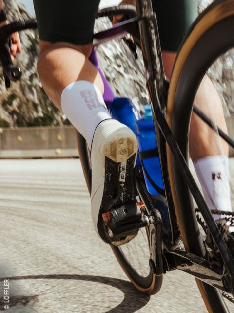 Primo piano del piede di un ciclista su un pedale, con scarpa da ciclismo bianca, borraccia blu e movimento sfocato su una strada.