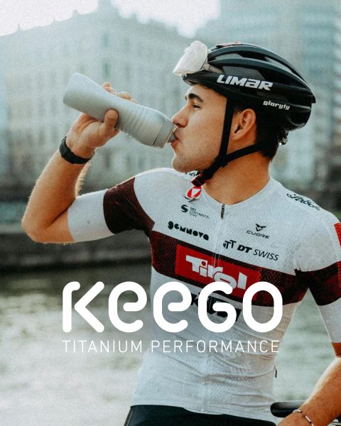 Un ciclista in una maglia bianca e rossa beve da una bottiglia e indossa un casco su cui è visualizzato il testo "KEEGO Titanium Performance".