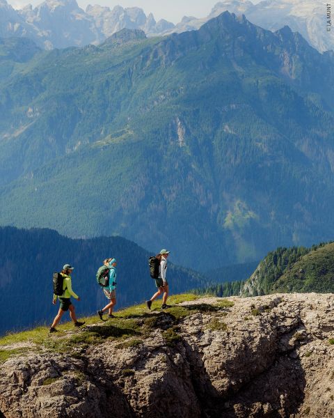 Tre escursionisti con zaini camminano lungo una cresta rocciosa con alte montagne boscose sullo sfondo sotto un cielo limpido.