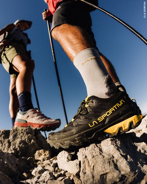 Primo piano di due escursionisti in piedi su un terreno roccioso, che guardano le loro scarpe da trail running La Sportiva sotto un cielo azzurro limpido.