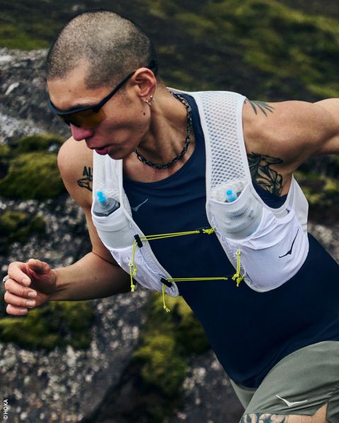 Una donna in abbigliamento sportivo e con un cappello indossa un gilet da corsa con borracce e si trova di lato davanti a un semplice sfondo bianco.