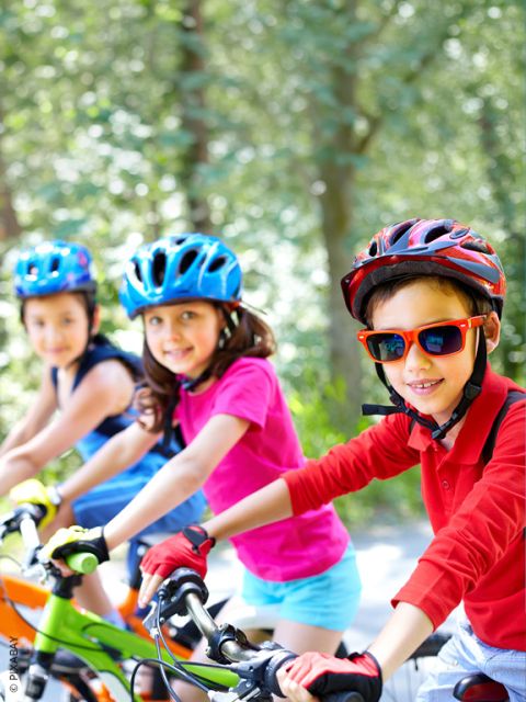 Tre bambini con i caschi vanno in bicicletta all'aperto in una giornata di sole, sorridendo alla telecamera. Lo sfondo è pieno di alberi verdi.
