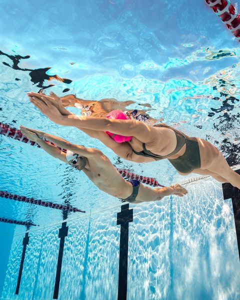 Due nuotatori, uno con una cuffia rosa e l'altro con una cuffia bianca, scivolano sott'acqua fianco a fianco in una piscina, nuotando in corsie separate.