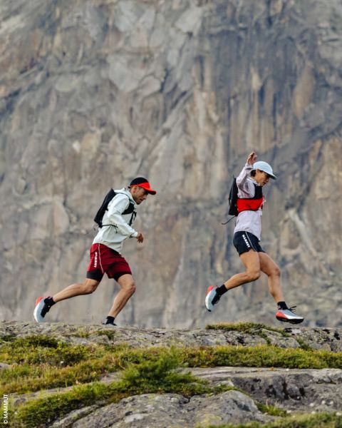 Due persone camminano su un sentiero roccioso davanti a una ripida scogliera di montagna, indossano cappelli, zaini e abbigliamento sportivo.