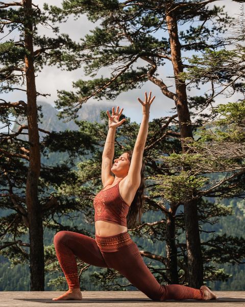 Una donna in abbigliamento sportivo rosso pratica yoga all'aperto su una terrazza di legno, circondata da alberi alti e montagne, e allunga le braccia sopra la testa in una posizione di affondo.
