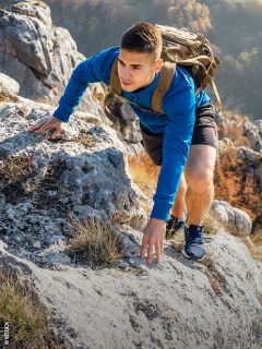 Un giovane uomo in una felpa blu e con uno zaino scala un versante roccioso di montagna, circondato da fogliame autunnale.