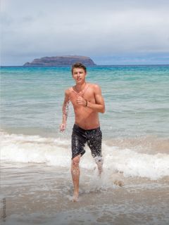Ein hemdsärmeliger Mann mit Tattoos und schwarzen Badeshorts steht bei Sonnenuntergang im flachen Meerwasser und schaut in die Kamera. Hinter ihm rollen die Wellen heran.