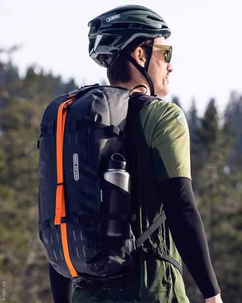 Una persona che indossa un casco verde e occhiali da sole, con uno zaino nero con una striscia arancione e una bottiglia d'acqua, si trova all'aperto in una zona boscosa.