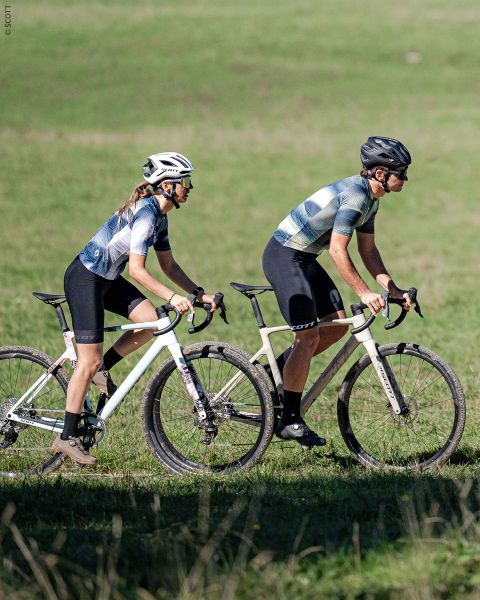 Due ciclisti con caschi e abbigliamento da ciclismo guidano biciclette bianche fianco a fianco su un sentiero erboso, con un prato verde sullo sfondo.