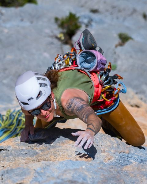 Un arrampicatore con casco e attrezzatura scala una parete rocciosa e tende le mani sporche di gesso verso l'alto sotto la luce splendente del sole.