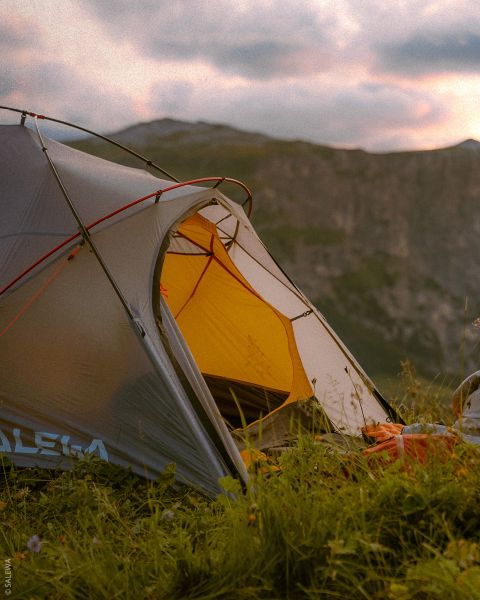 Una tenda grigia e gialla si trova su un prato con montagne sullo sfondo sotto un cielo nuvoloso al tramonto.