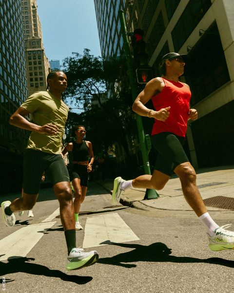 Tre persone in abbigliamento sportivo e scarpe da ginnastica camminano su una strada in città, con alti edifici e la luce del sole sullo sfondo.