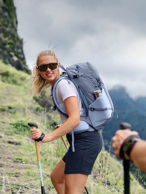 Una donna con occhiali da sole cammina lungo un sentiero di montagna erboso con bastoncini da trekking e uno zaino grigio, sorridendo alla telecamera.