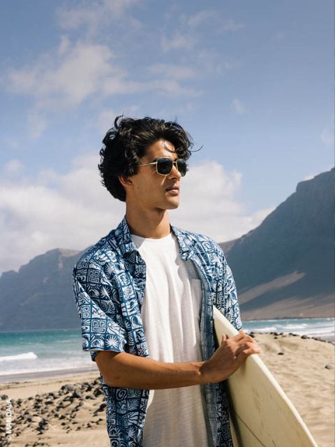 Uomo con occhiali da sole e camicia a motivi blu tiene in mano una tavola da surf e sta in piedi su una spiaggia sabbiosa con montagne e mare sullo sfondo.