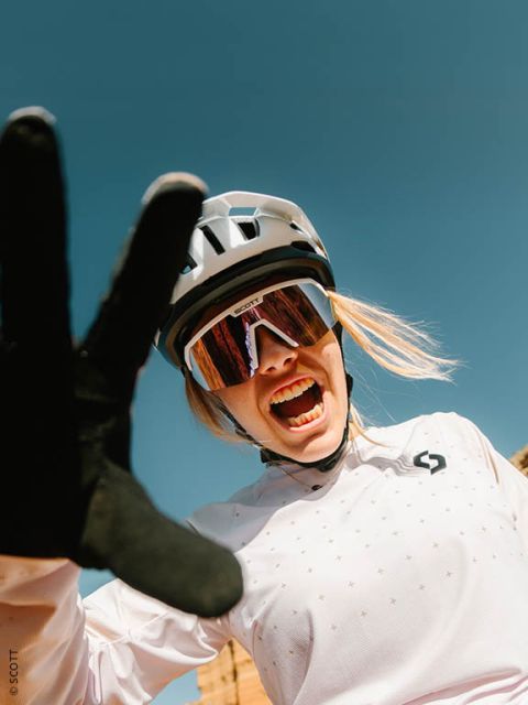 Un ciclista sorridente con casco, occhiali da sole e guanti si allunga verso la telecamera davanti a un cielo azzurro brillante.