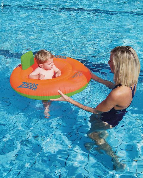 Una donna tiene in braccio un bambino che è seduto in una ciambella da nuoto arancione e verde in una piscina.